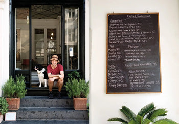 A man in a brown hat and red shirt sits on steps beside a black and white dog, smiling. Behind them is a glass door. To the right, an Apartamento Atelier September: A Place for Daytime Cooking chalkboard menu is visible.