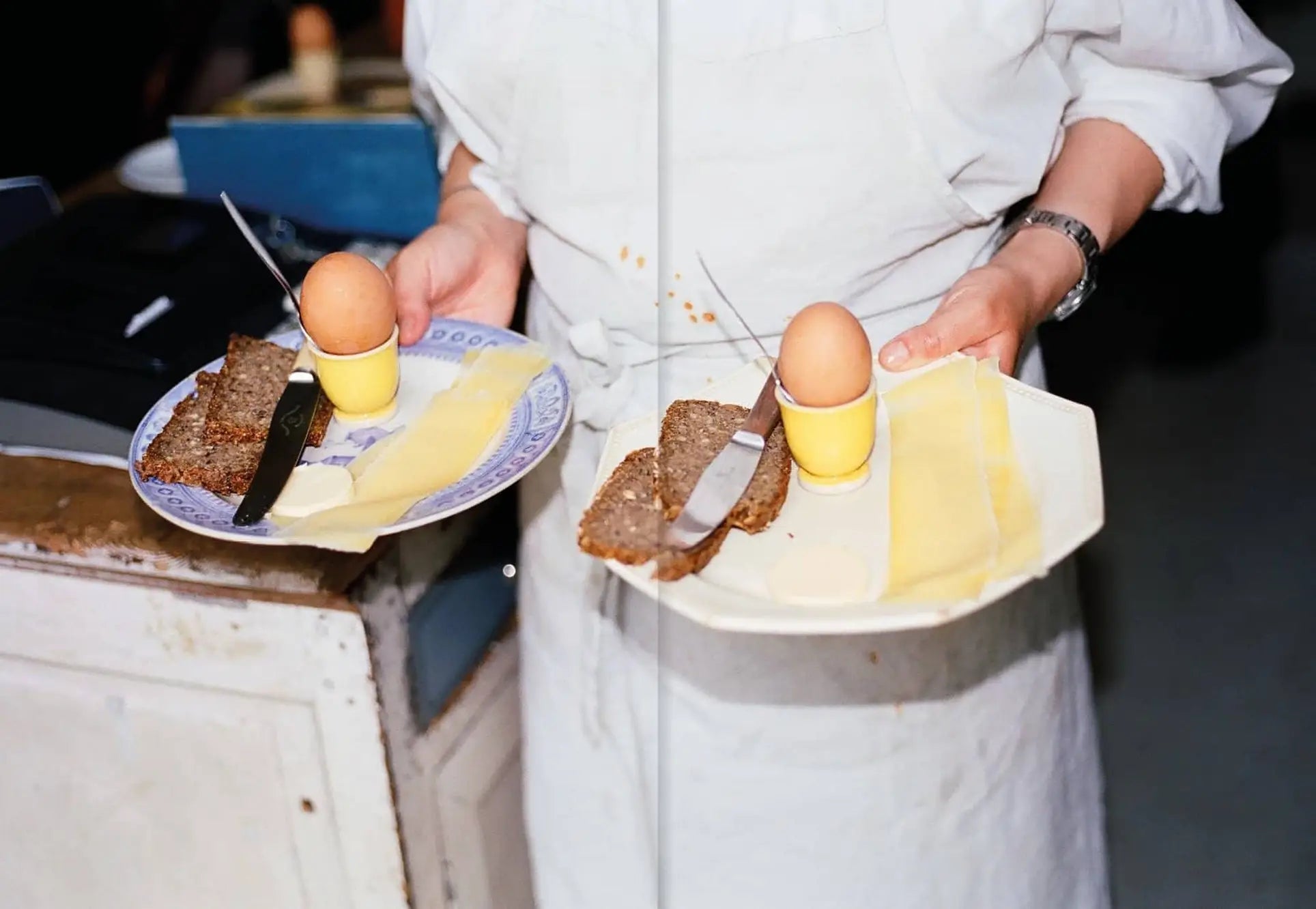 A person in a white apron holds two plates with boiled eggs, cheese, bread, and butter knives—an inviting scene inspired by recipes from Apartamento Atelier September: A Place for Daytime Cooking by Apartamento.