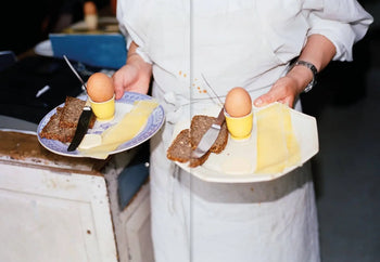 A person in a white apron holds two plates with boiled eggs, cheese, bread, and butter knives—an inviting scene inspired by recipes from Apartamento Atelier September: A Place for Daytime Cooking by Apartamento.