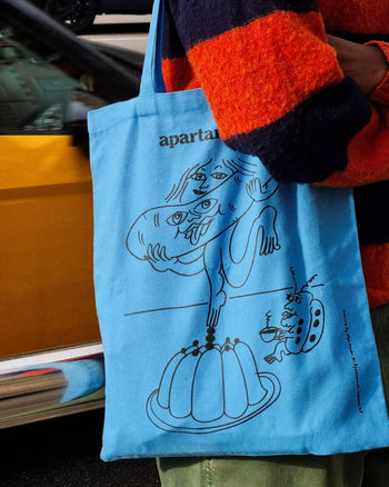 Someone in an orange and navy striped sweater holds the Apartamento Cookbook #8 Tote Bag by Apartamento, featuring a whimsical black line drawing, abstract figures, and a playful jelly mold—an artistic, sustainable accessory.