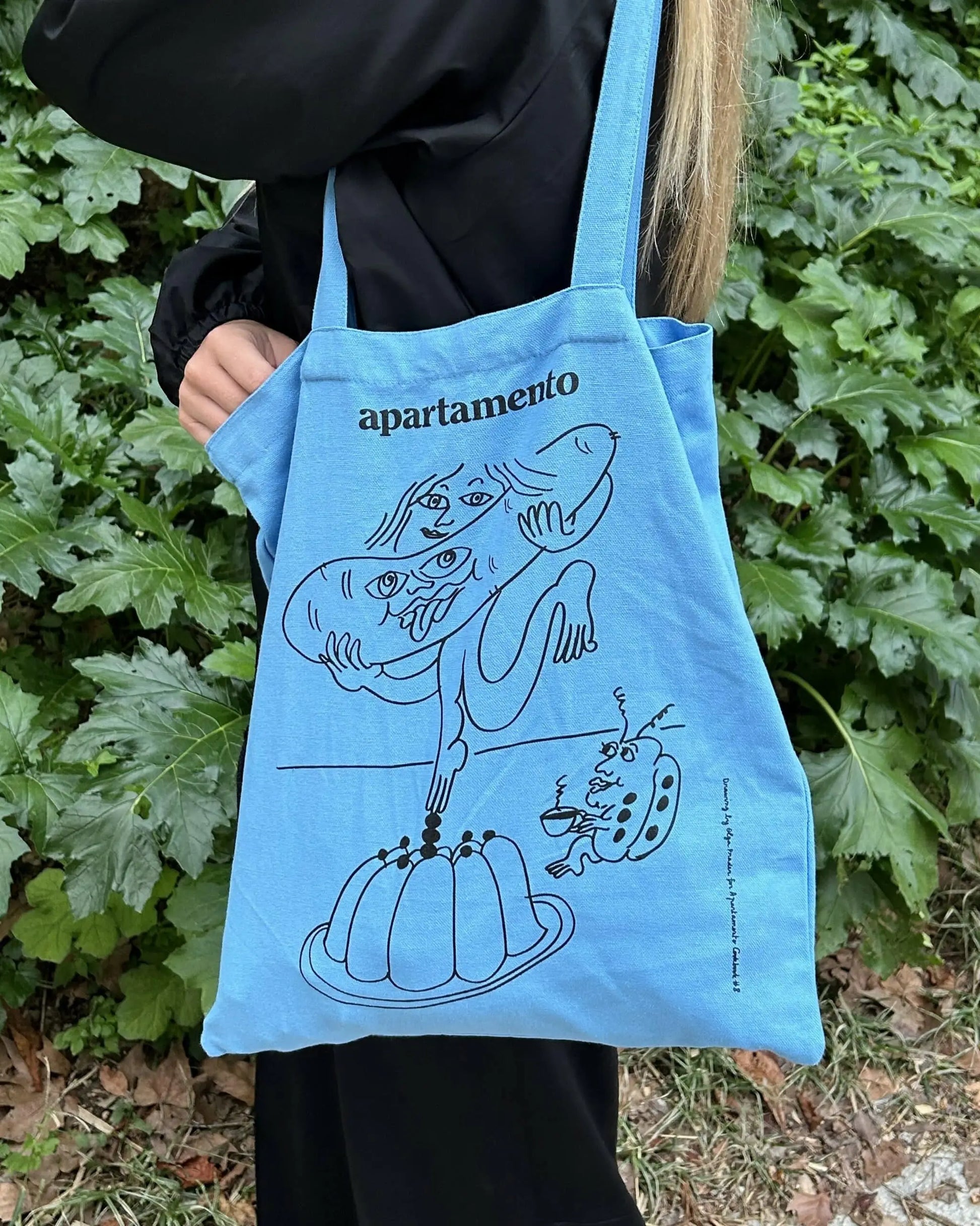 A person holds the Apartamento Cookbook #8 Tote Bag by Apartamento, a blue bag adorned with a playful black line drawing of a bread-eating figure. Green leafy plants in the background evoke a fresh, sustainable vibe.