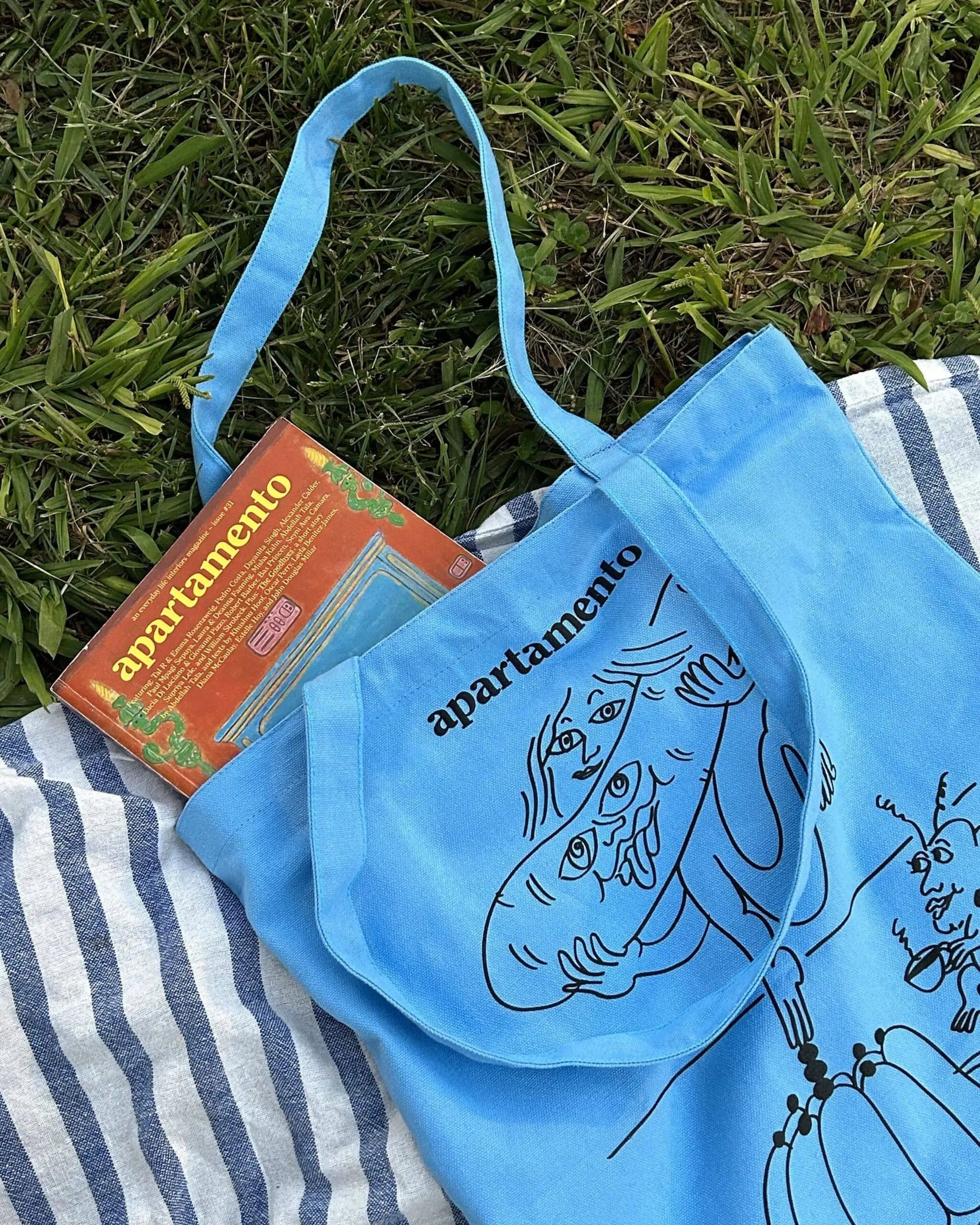 The Apartamento Cookbook #8 Tote Bag by Apartamento, featuring blue abstract line art and the word apartamento, rests on a striped blanket with an Apartamento Cookbook peeking out, chicly hinting at style and sustainability.