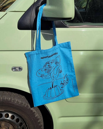 The Apartamento Cookbook #8 Tote Bag by Apartamento, featuring a quirky black line illustration and branding, hangs on the side mirror of a light green car. The design depicts a character enjoying a jelly dessert.