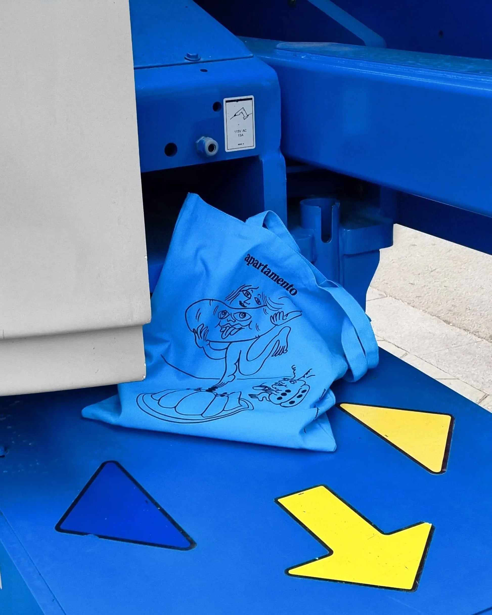 The Apartamento Cookbook #8 Tote Bag by Apartamento, featuring a cartoon character and bold lettering, sits on a blue machine with colorful arrows—a playful nod to the bags stylish and sustainable design.