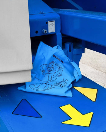 The Apartamento Cookbook #8 Tote Bag by Apartamento, featuring a cartoon character and bold lettering, sits on a blue machine with colorful arrows—a playful nod to the bags stylish and sustainable design.