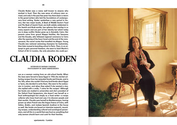 A woman with long hair sits on a wooden chair in a sunlit room at Siwa Oasis, holding a flower—a serene scene from Spring/Summer 2022, captured in Apartamento Magazine Issue #29 by Apartamento. Lush plants flourish outside the window.