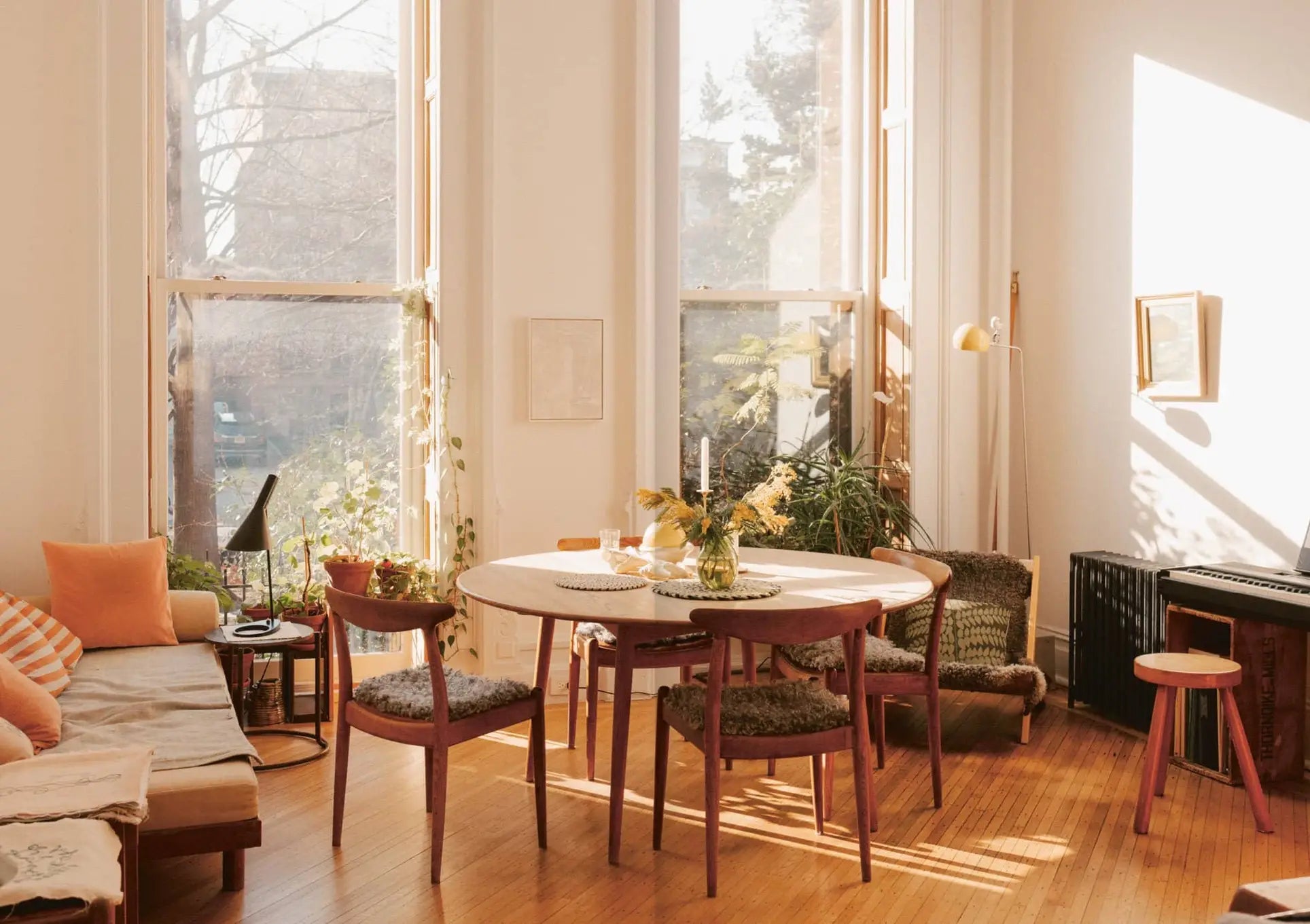 Bright, sunlit dining room with large windows, a round table and chairs, plants, piano, and cozy seating on warm wood floors. Natural light fills the inviting space—perfect for reading Apartamento Magazine Issue #29 by Apartamento.