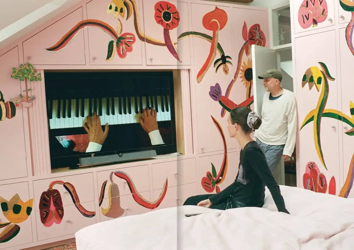 A woman sits on a bed watching TV as a man stands nearby; the room’s pink walls and cabinets feature whimsical, colorful flowers inspired by Tal R’s Spring Summer 2023 aesthetic, as seen in Apartamento Magazine Issue #31 by Apartamento.