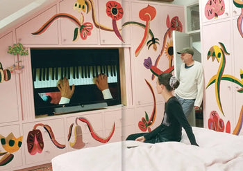 A woman sits on a bed watching TV as a man stands nearby; the room’s pink walls and cabinets feature whimsical, colorful flowers inspired by Tal R’s Spring Summer 2023 aesthetic, as seen in Apartamento Magazine Issue #31 by Apartamento.