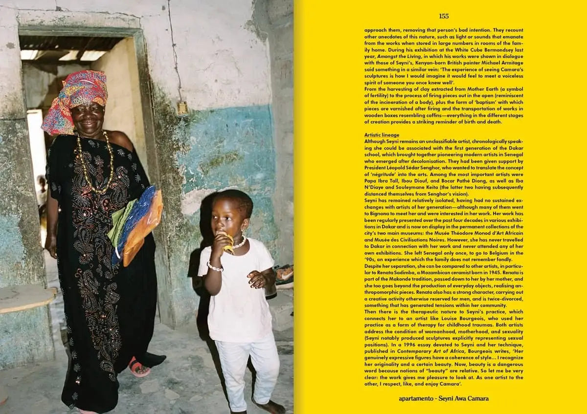 An elderly woman in vibrant traditional attire stands by a child indoors near a faded blue and white wall, sunlight streaming in. Inspired by Apartamento Magazine Issue #31 from Apartamento, Spring Summer 2023.