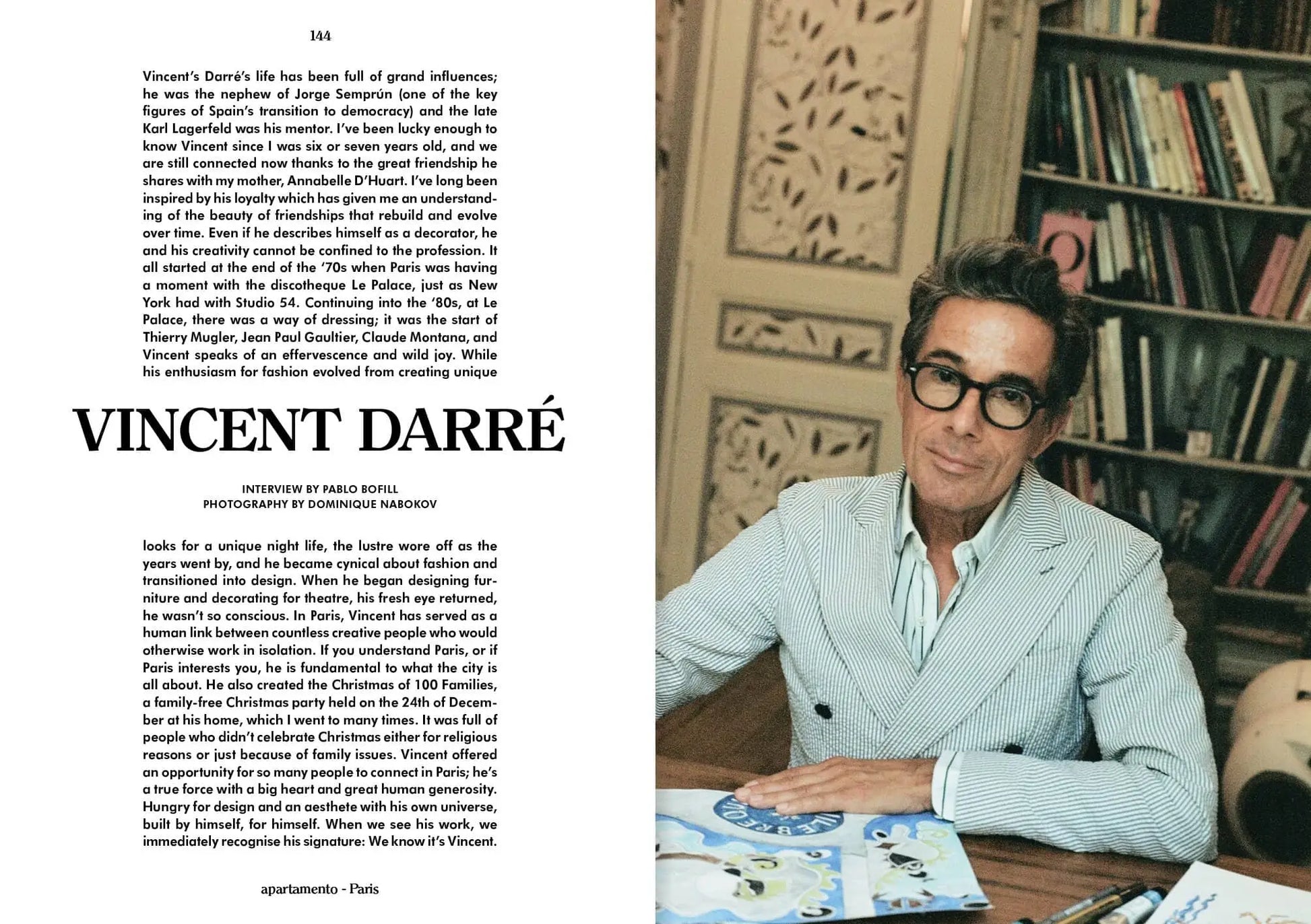 A man with dark hair and glasses, wearing an Autumn/Winter 2023-24-inspired blazer, sits at a desk with art supplies. Apartamento Magazine Issue #32 by Apartamento is on the shelf amid books and a decorative panel behind him.