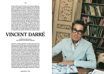 A man with dark hair and glasses, wearing an Autumn/Winter 2023-24-inspired blazer, sits at a desk with art supplies. Apartamento Magazine Issue #32 by Apartamento is on the shelf amid books and a decorative panel behind him.