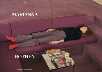 On a purple sectional, someone in a black top, mauve pants, and red socks reclines—a scene echoing Kazuo Shinohara’s minimalism. Nearby rests Apartamento Magazine Issue #32 by Apartamento on a low table with books, a plate, and a cup.