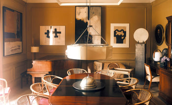 A stylish dining room features a dark wood table, rattan chairs, abstract art on yellow walls, a grand piano, and a modern chandelier inspired by Apartamento’s Miguel Milá: A Life in Design. Natural light fills the space.