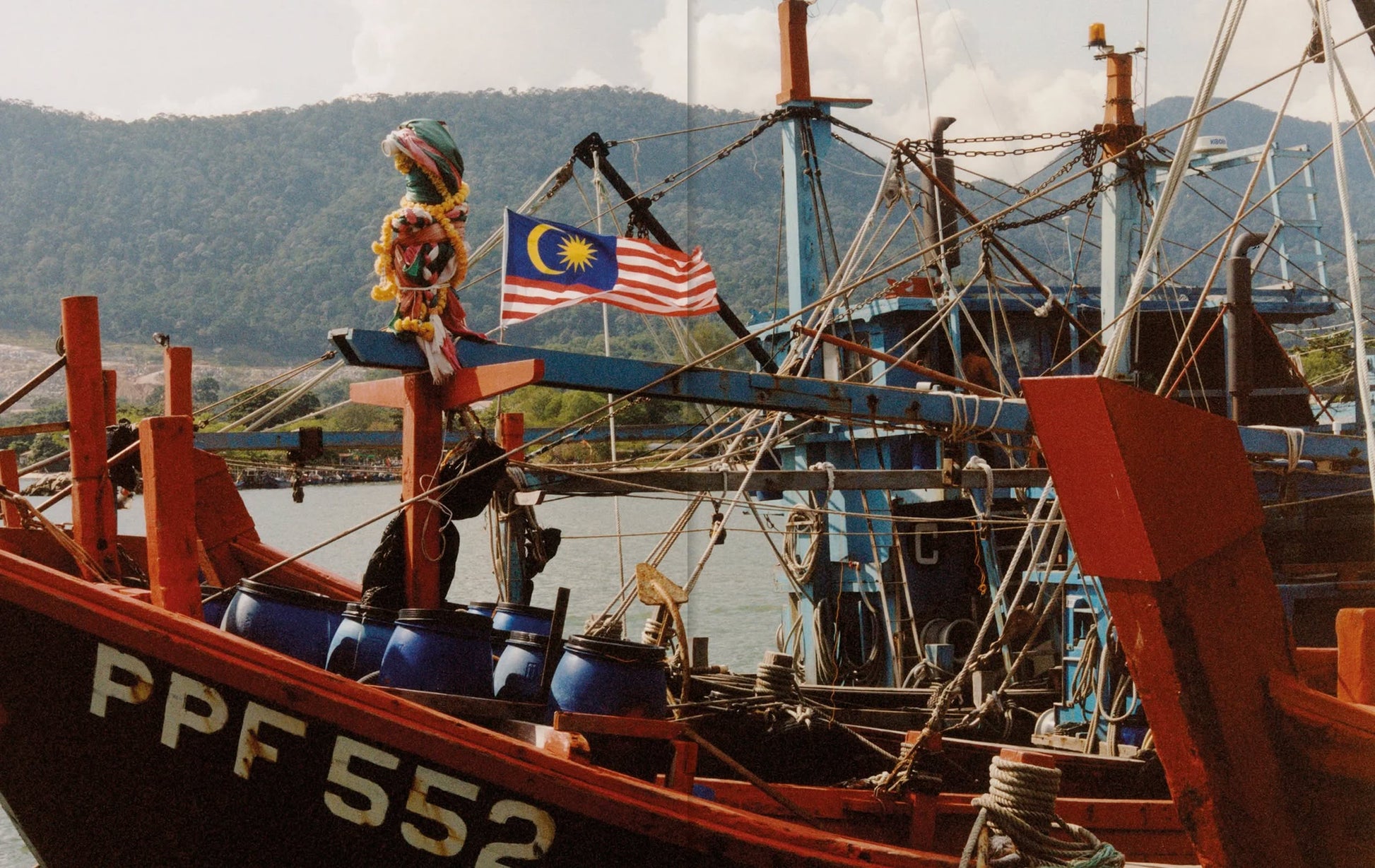 A red fishing boat marked PPF 552, with blue barrels and a Malaysian flag, is docked—ready to supply Penangs cuisine, as featured in Apartamento PENANG: Recipes & Wanderings Around an Island in Malaysia by Apartamento. Other boats and green hills are behind.
