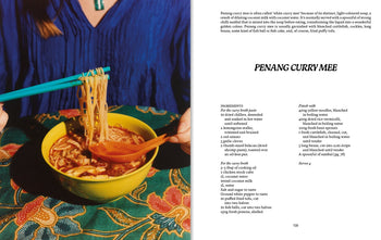 A person lifts noodles with orange chopsticks from a bowl of Penang curry mee, set atop a patterned table, as the recipe appears on the right page of Apartamento PENANG: Recipes & Wanderings Around an Island in Malaysia by Apartamento.