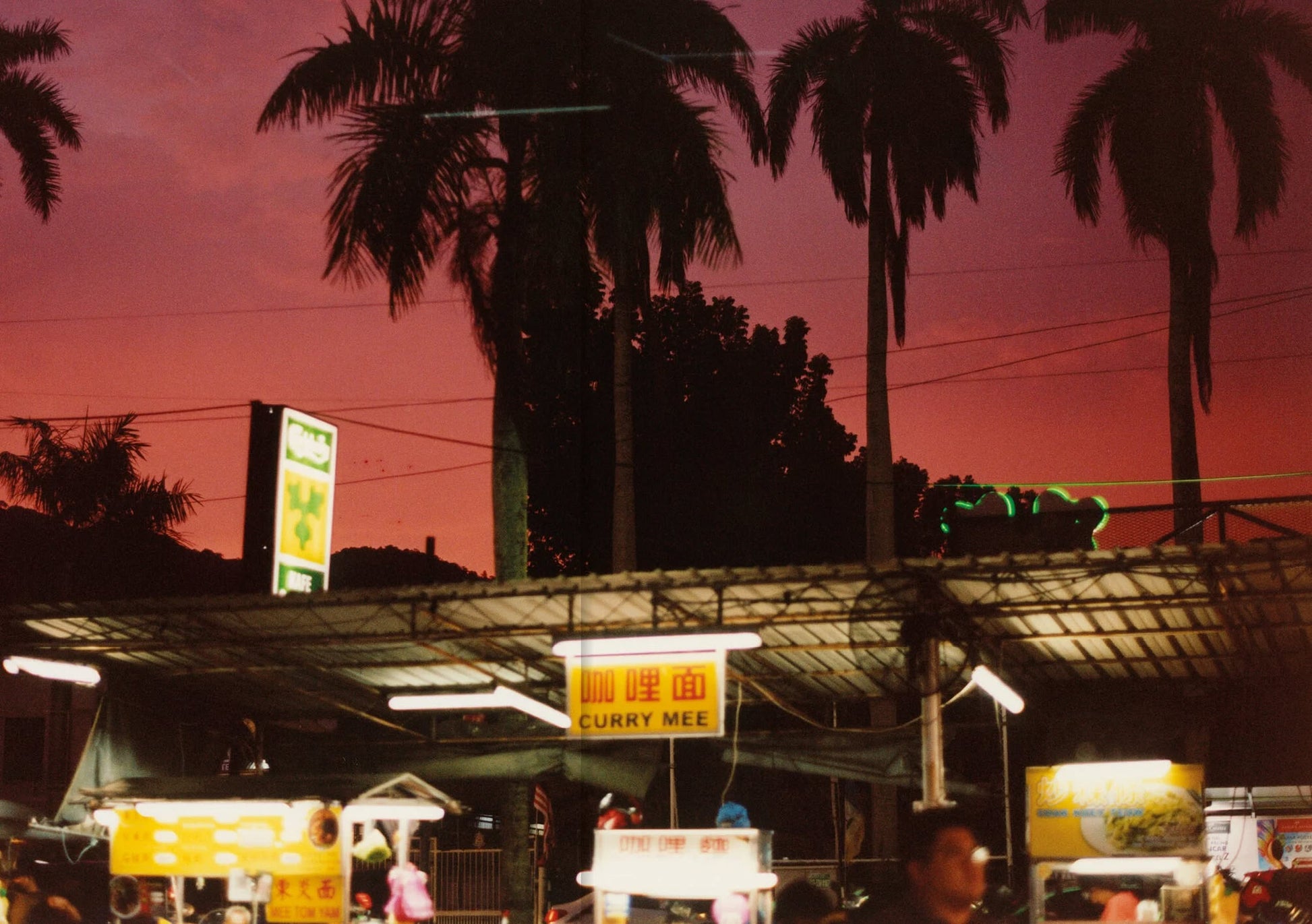As dusk falls, people gather by a Curry Mee food stall under palm trees and a vivid sunset, echoing the culinary adventure found in Apartamento PENANG: Recipes & Wanderings Around an Island in Malaysia by Apartamento.