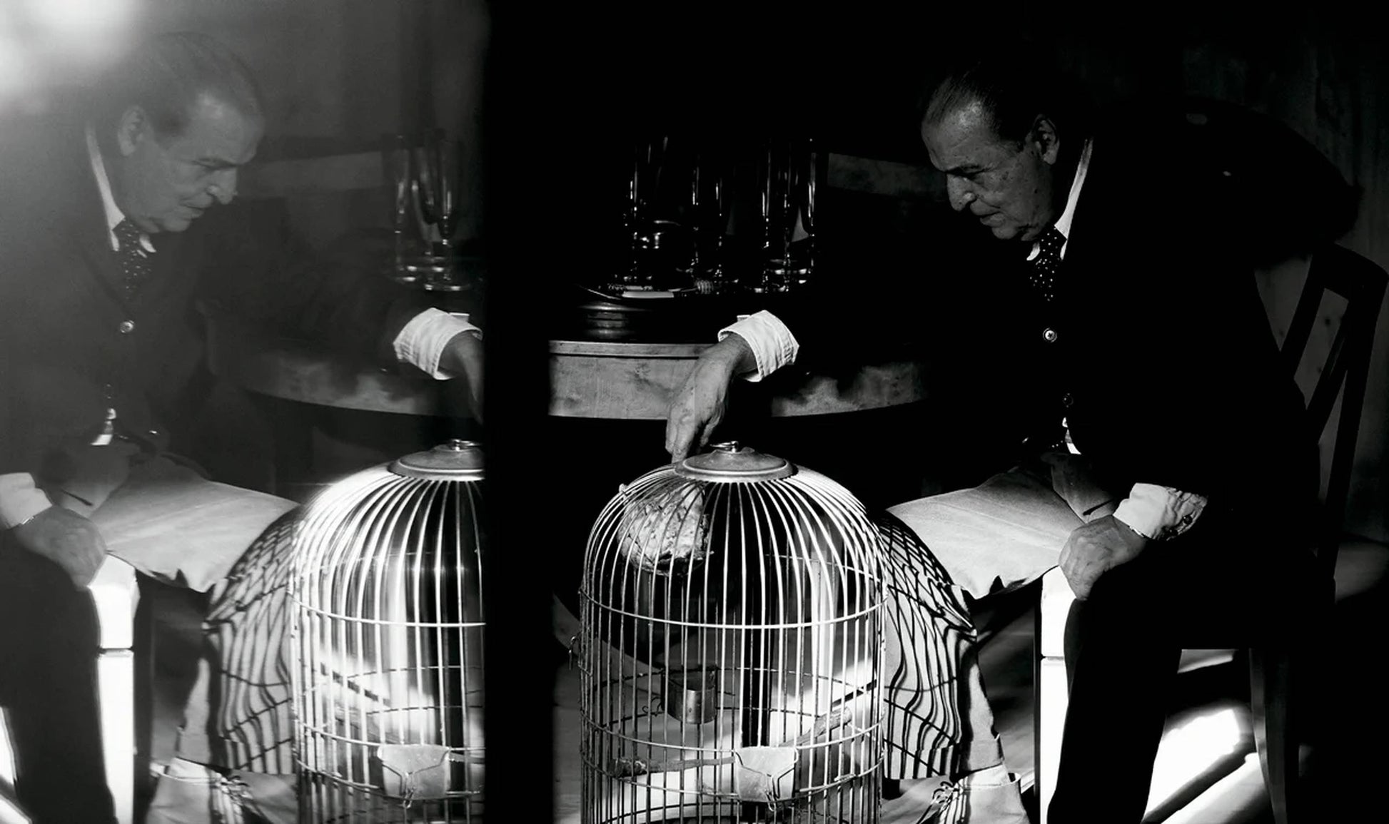 A black-and-white photo from Aptamento The House of Xavier Corberó by Apartamento shows a man in a suit at his Esplugues de Llobregat home, leaning on a birdcage. The dim light and mood evoke surrealist sculpture.
