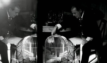 A black-and-white photo from Aptamento The House of Xavier Corberó by Apartamento shows a man in a suit at his Esplugues de Llobregat home, leaning on a birdcage. The dim light and mood evoke surrealist sculpture.