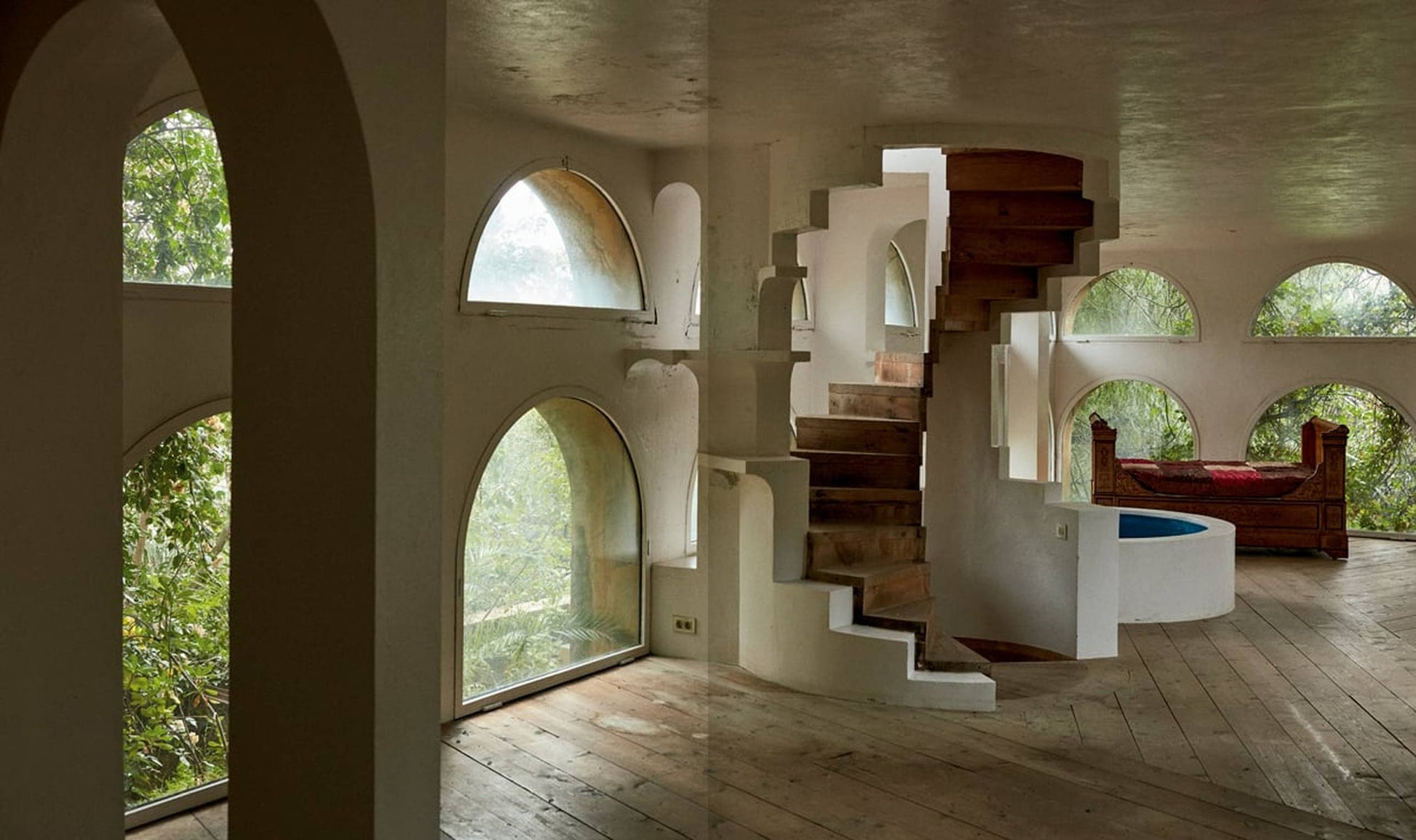 A sunlit room with arched windows, rustic wood floors, and a spiral staircase channels the surrealist style of Apartamento The House of Xavier Corberó by Apartamento, featuring a cozy bed nestled amid greenery outside.