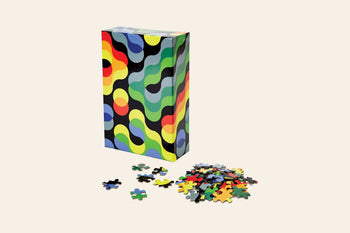The Areaware Dusen Dusen - Pattern Puzzle, a 500-piece jigsaw with an abstract wavy design, stands upright beside scattered pieces on a plain background—ideal for design enthusiasts.