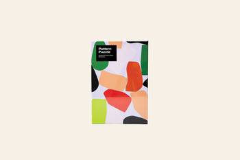 The Areaware Dusen Dusen - Pattern Puzzle, a 500-piece jigsaw with an abstract, colorful design on white, comes in a rectangular box from Areaware and sits centered on a beige surface—ideal for design lovers.