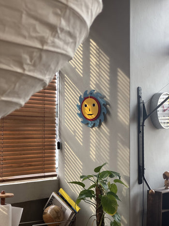 An Areaware Everybody Clock with a blue and yellow sun-shaped design by Areaware hangs on the wall, casting shadows from window blinds. Below, a green potted plant stands near books and a globe while a paper lantern brightens the foreground.