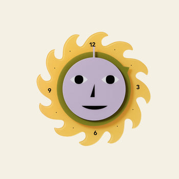 The Areaware Everybody Clock by Areaware is a sun-shaped wall clock with a yellow flame-like border and a purple face featuring simple black eyes, nose, and mouth. Numbers 3, 6, 9, and 12 are marked around the edge for easy time reading.