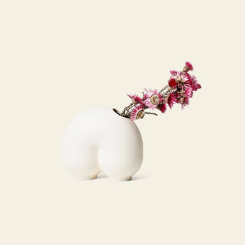The Areaware Kirby Vase Enny by Areaware, a white ceramic vase inspired by human curves, displays vibrant pink thistle flowers—an attention-grabbing piece ideal for creative floral arrangements on a minimalist backdrop.
