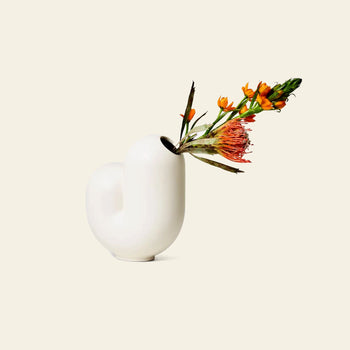The Areaware Kirby Vase Jay by Areaware is a white, curvy ceramic vase with a modern design that holds orange and pink flowers, making it perfect for simple floral arrangements. The vase pops against a plain, light beige background.