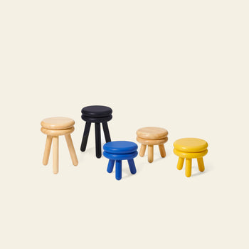 Five Areaware Pluma Stool Short stools by Areaware, each with three legs, are grouped on a beige background. The stools feature contemporary design and come in light wood, black, blue, yellow, and natural wood finishes.