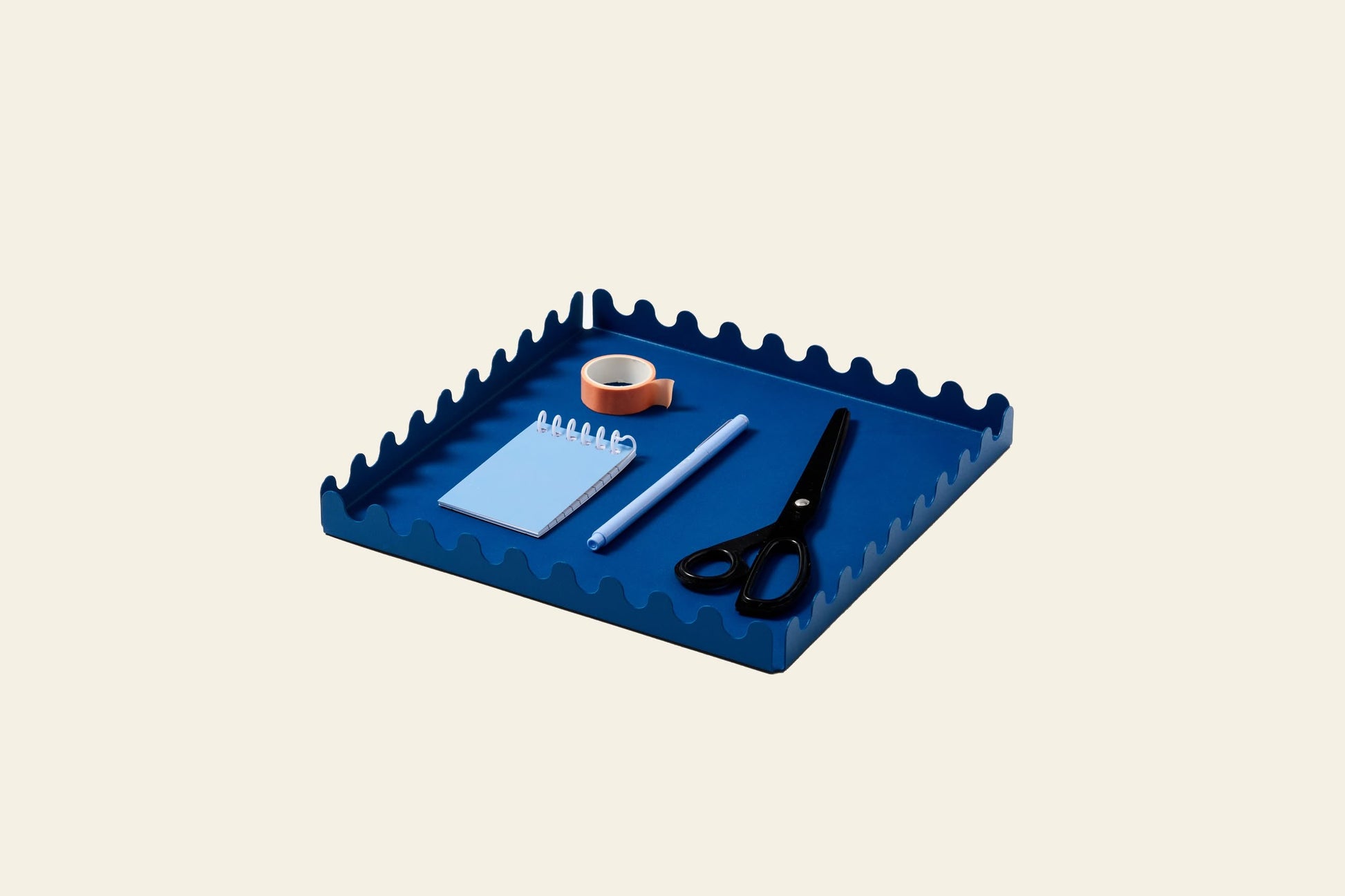 The Areaware Scape Tray in blue, with wavy edges, displays a notepad, tape roll, pen, and black scissors—all neatly arranged on a light beige background for a minimalist look.