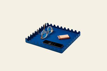 An Areaware Scape Tray by Areaware, featuring a blue wavy-edged minimalist design, holds round eyeglasses, a pink rectangular case, and a slim black remote control against a plain light background.