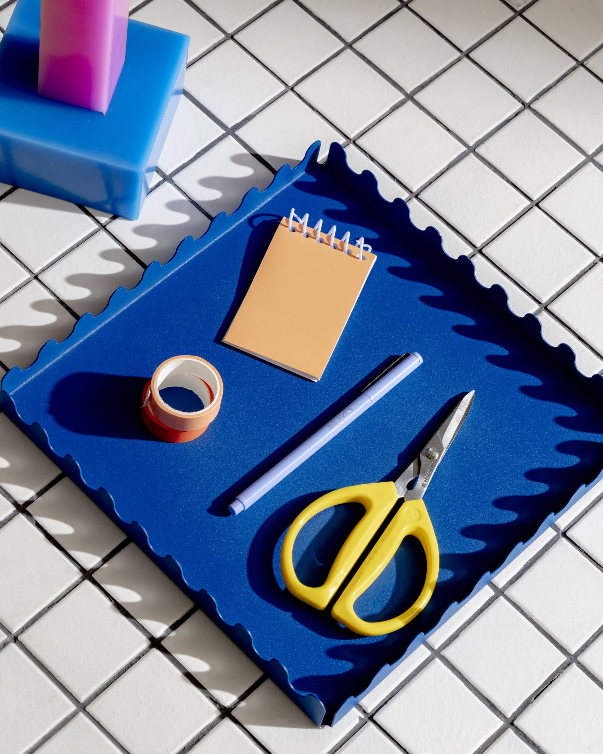 An Areaware Scape Tray by Areaware with wavy edges sits on white tile, holding yellow-handled scissors, a blue pen, a spiral notepad, and tape. A pink and blue item appears at the corner.