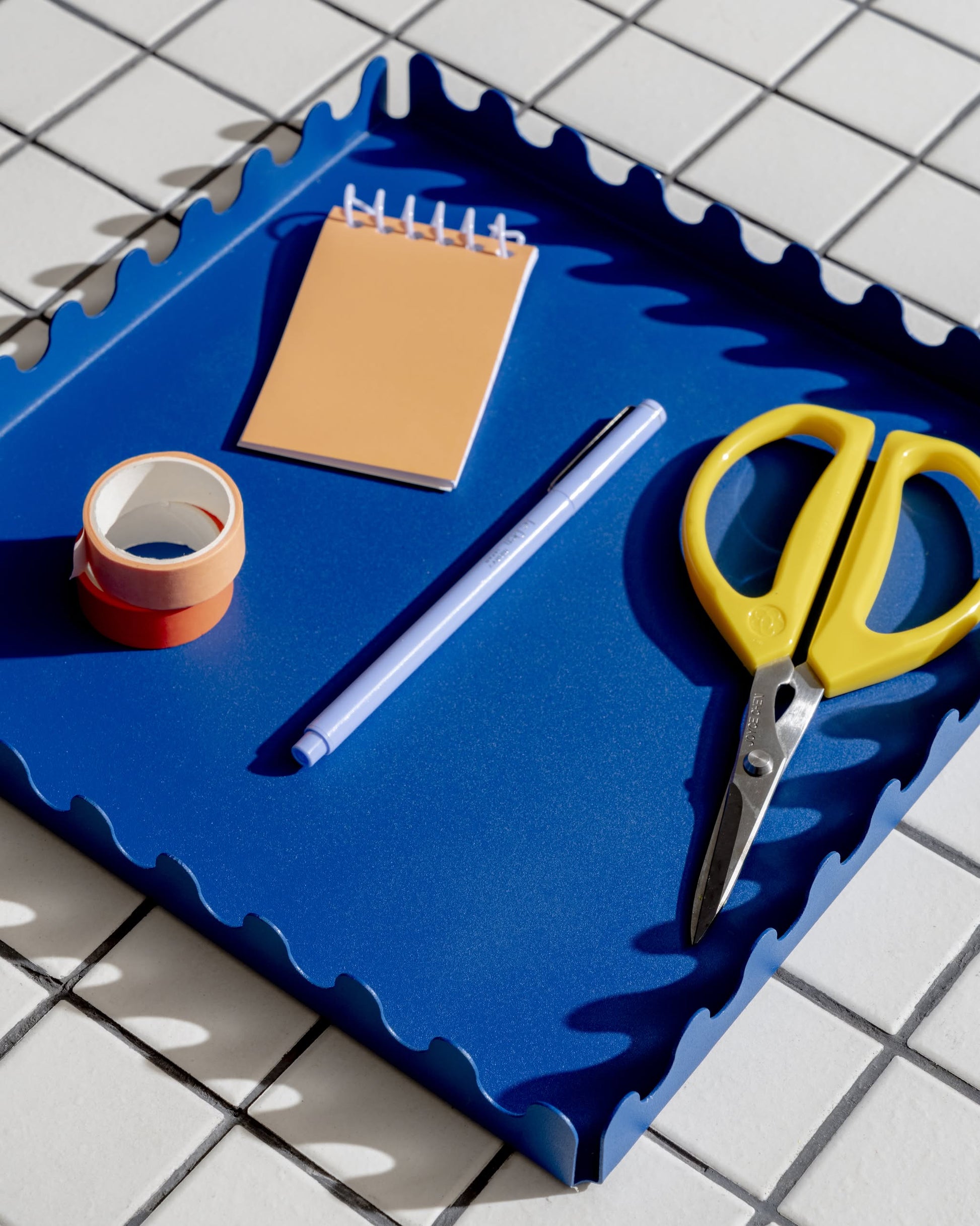 A blue Areaware Scape Tray with a minimalist design holds a peach notepad, purple pen, red and white tape rolls, and yellow scissors. The tray by Areaware rests on white tiles with dark grid lines.