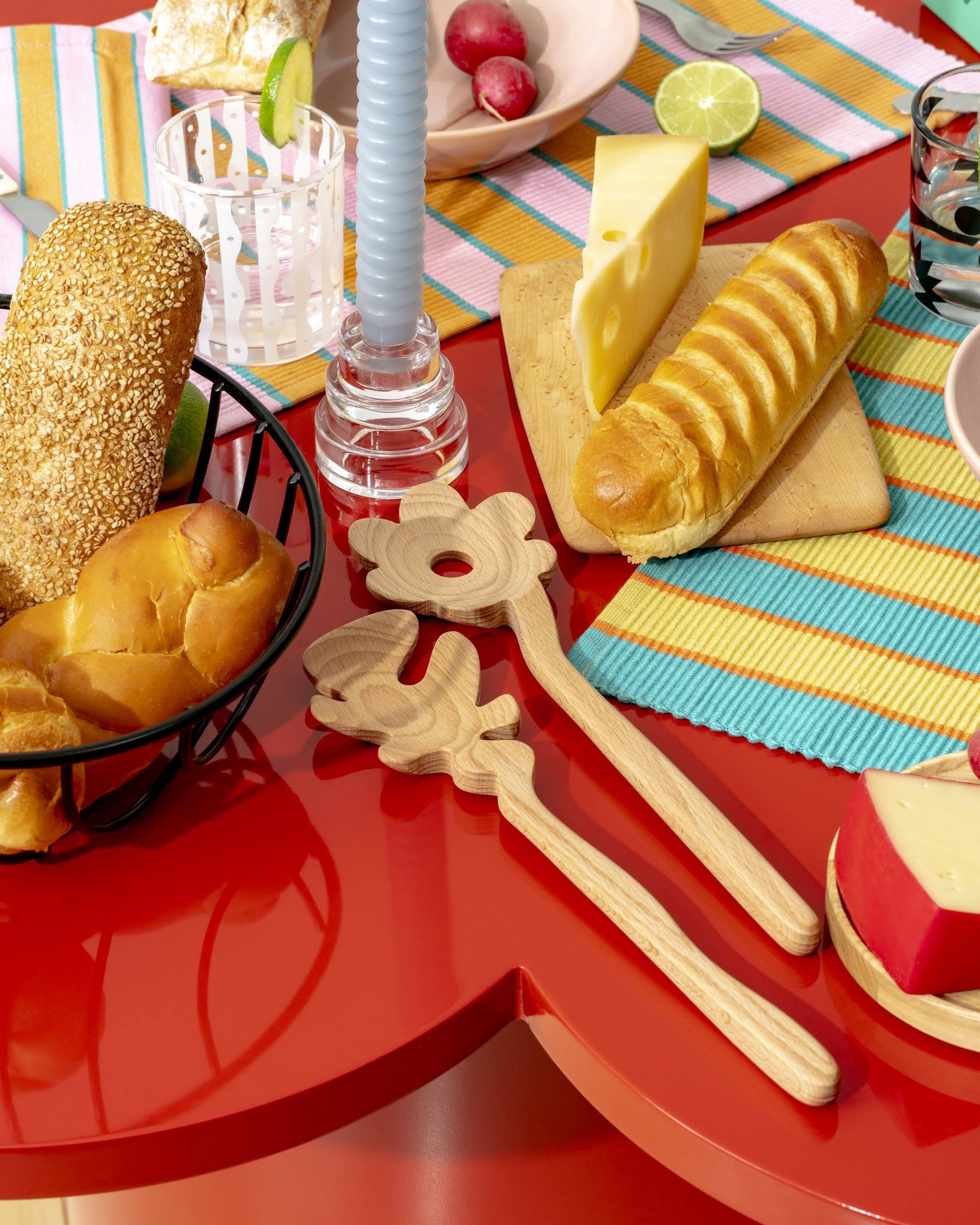 The Areaware Serving Friends - Flowers by Areaware displays charming utensils, wooden spoons, a basket of bread, sliced cheese, a striped napkin, glass of water, blue spiral candle, and assorted food for a cozy meal.
