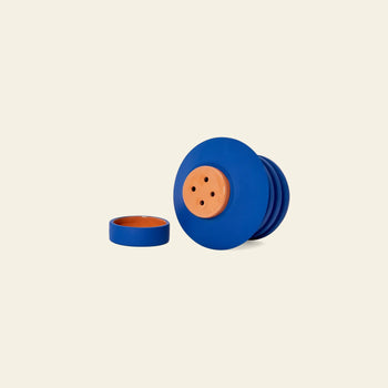 The Areaware Strata Plant Vessel by Areaware is a blue and terracotta clay container with a sculptural design and four holes on the lid, shown against an off-white background with the lid placed beside the base.