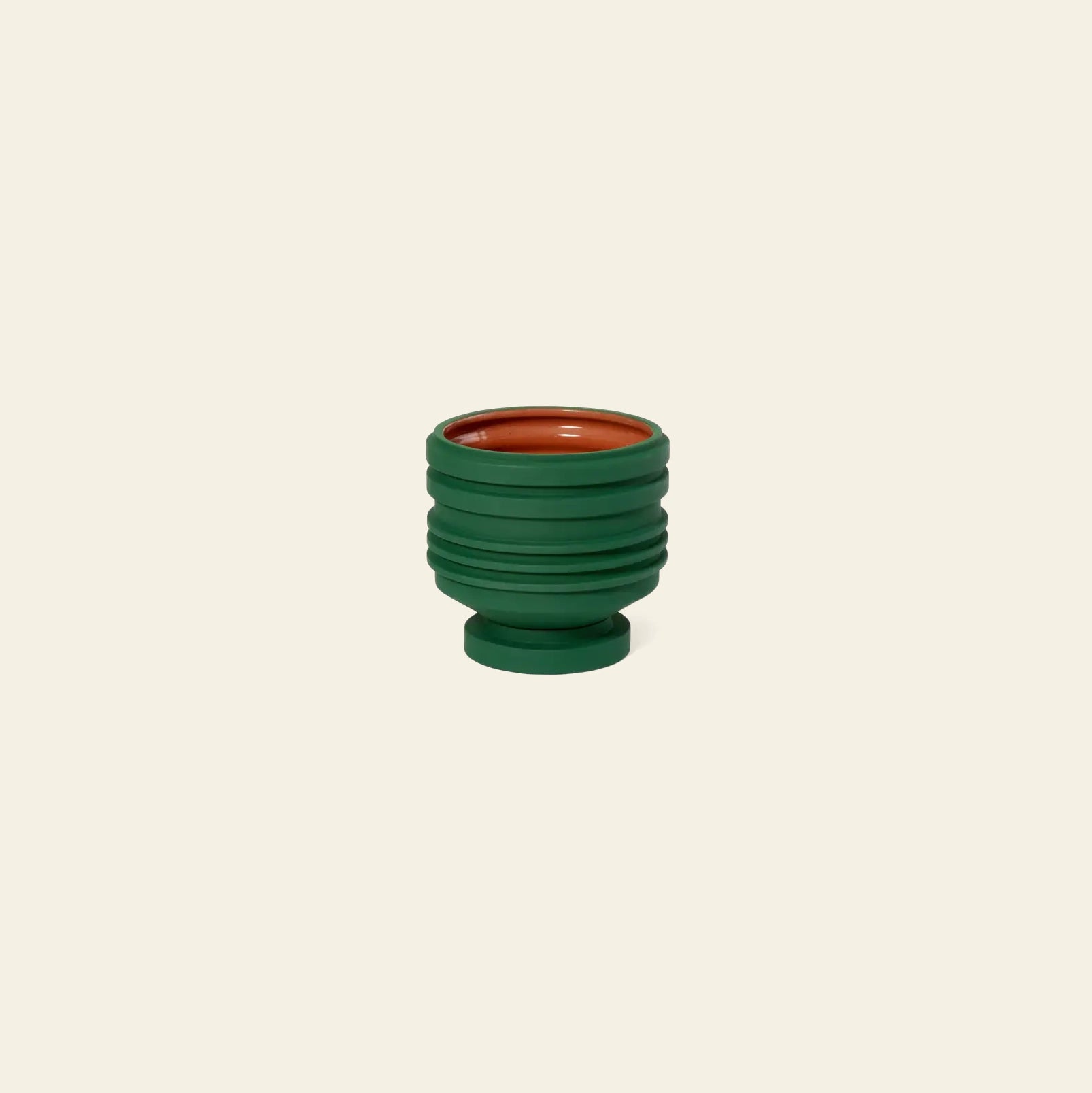 The Areaware Strata Plant Vessel by Areaware is a small, round ceramic planter with sculptural horizontal ridges, dark green exterior, and terracotta orange interior, inspired by Simone Brewster and shown against a plain beige background.