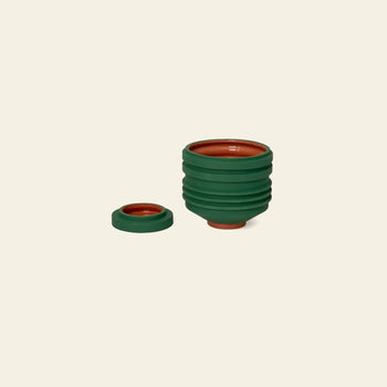 The Areaware Strata Plant Vessel by Areaware, with a sculptural design, green exterior, and orange interior, is displayed expanded on a light background with its green lid set beside it.