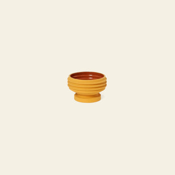 The Areaware Strata Plant Vessel by Areaware features a sculptural design with a mustard yellow ribbed exterior and brown interior, set on a matching pedestal, reminiscent of Simone Brewster’s style.
