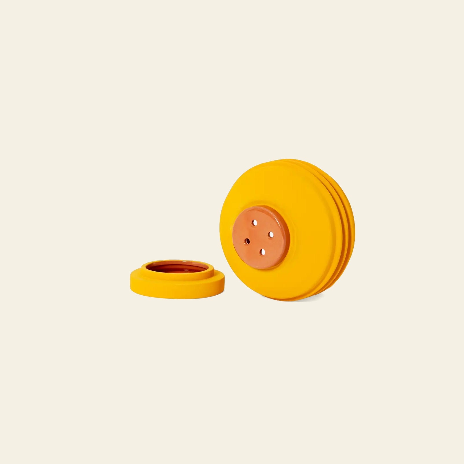 A round, yellow plastic object with a copper center and four holes—the Areaware Strata Plant Vessel by Areaware—has its lid removed and placed beside it on a plain, light background, reflecting Simone Brewster’s sculptural ceramics style.