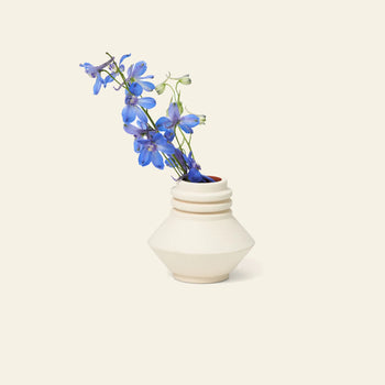 The Areaware Strata Vase Mini by Areaware, a small geometric white ceramic vase, holds vibrant blue flowers against a plain beige background, evoking sculptural designs reminiscent of Simone Brewster.