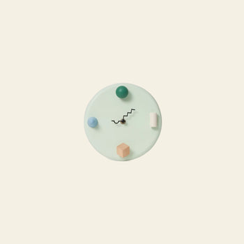 The Areaware Wall Clock by Areaware features a minimalist pale circular face with abstract 3D geometric markers in blue, green, peach, and white, plus black zigzag hands, all set against a plain beige background.