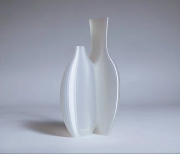 Two tall, modern, semi-translucent white vases of varying heights stand side by side on a light grey surface—a striking Argot Studio Double Vessel that reimagines classic form.