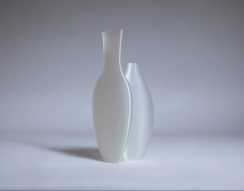 The Argot Studio Double Vessel by Argot Studio features two elegant matte white ceramic vases with elongated, curved forms—a modern take on classic shapes. One vase stands slightly taller and slimmer than the other, set against a soft gray background.