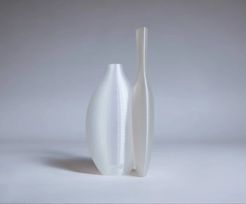 The Argot Studio Double Vessel by Argot Studio features two tall, modern white vases with smooth, minimalist forms, side by side on a light surface against a neutral gray background.