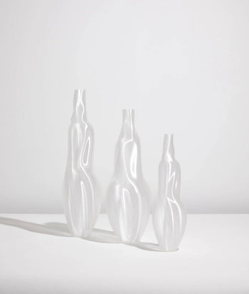 Three Argot Studio Éson Vessels (Small) by Argot Studio, with tall white sculptural forms and organic fluid silhouettes, stand side by side on a white surface against a plain background, casting soft left shadows—a modern take on classic design.
