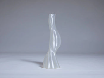 The Argot Studio Les Hortenses Vase, Small—tall, translucent, and abstract with a twisted silhouette—stands on a smooth light gray surface, offering a contemporary touch against a plain gray background.