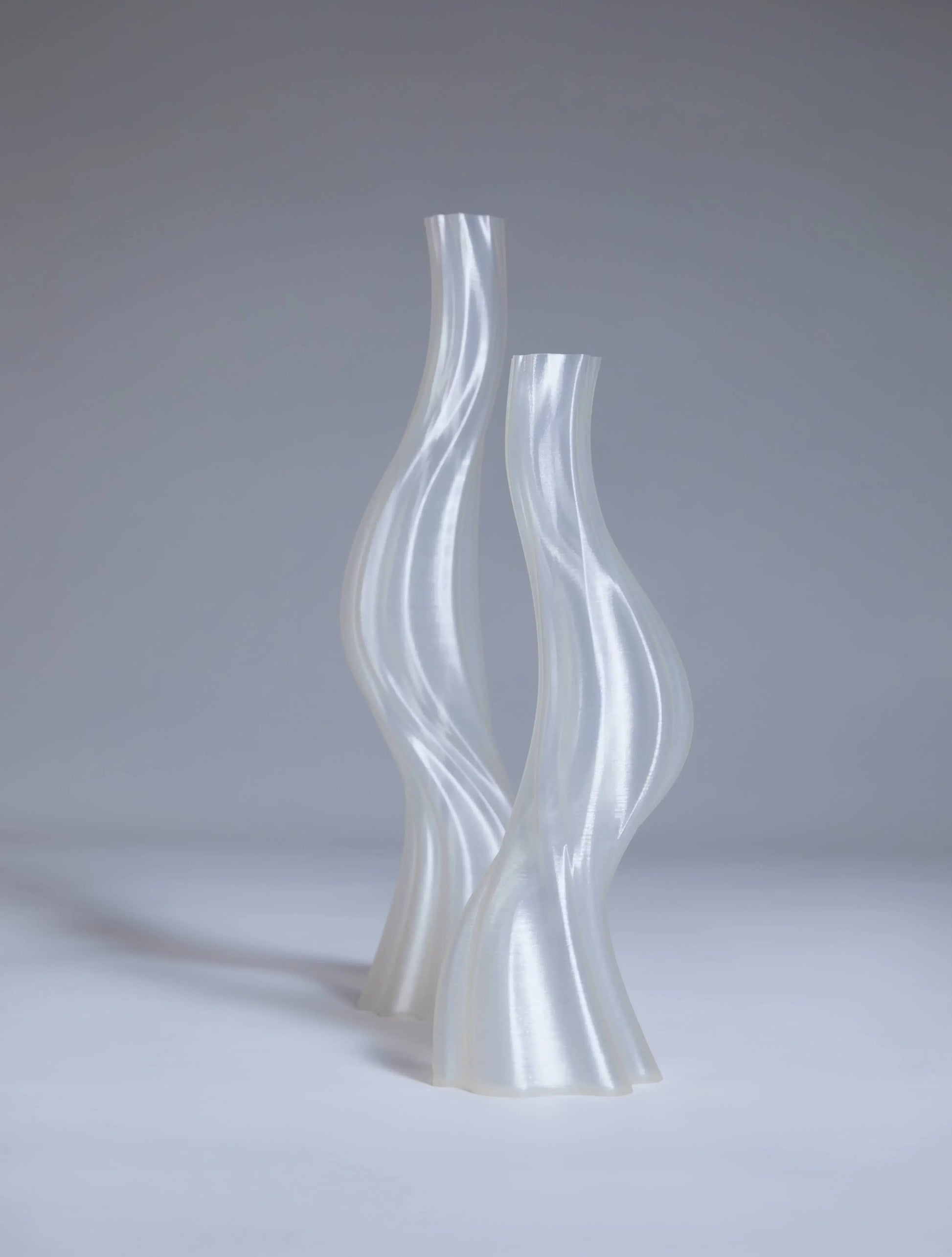Two tall, translucent vases—Argot Studio Les Hortenses Vases (Small) by Argot Studio—stand side by side on a neutral gray surface, showcasing a glossy, contemporary reinterpretation made from 3D printed material.