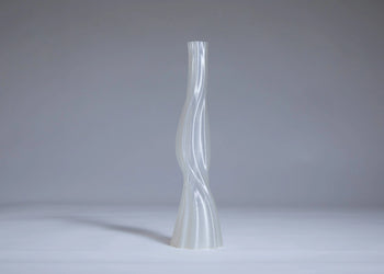 The Argot Studio Les Hortenses Vase, Tall, by Argot Studio features a slender, translucent contemporary form with a twisted sculptural design, standing on a light gray surface against a plain gray background.